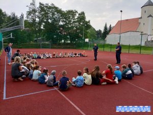 Grupa dzieci siedzi na boisku. Przed nimi stoi dwóch policjantów