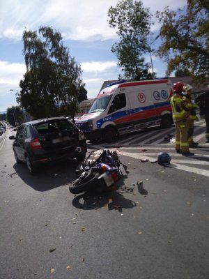 Na zdjęciu widać leżący motocykl i rozbity samochód osobowy. Obok stoi karetka pogotowia oraz strażacy