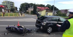 Na zdjęciu widać leżący motocykl i rozbity samochód osobowy. Obok stoi policjant ruchu drogowego oraz strażak