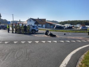 Zdjęcie przedstawia miejsce zdarzenia drogowego. Na drodze leży motocykl, przy pojeździe stoją strażacy