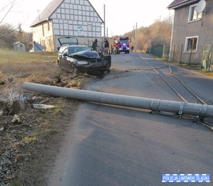 Zdjęcie przedstawia rozbity samochód BMW koloru ciemnego. Z tyłu widać straż pożarną