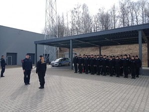 Grupa uczniów stoi na placu. Jedna z osób składa policjantowi meldunek