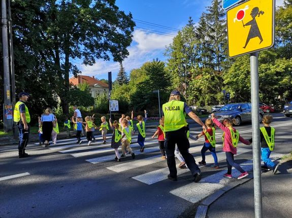 Dzieci przechodzą przez przejście dla pieszych. Policjant stoi na drodze. Policjantka idzie z dziećmi