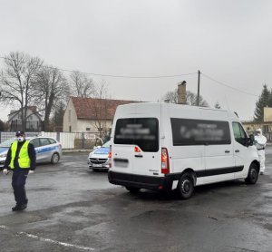 Na zdjęciu po lewej stronie widać dwa stojące radiowozy oraz policjanta w maseczce. Po prawej stronie widać duży biały bus, który jest kontrolowany przez policjanta ubranego w biały ochronny kombinezon