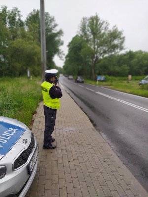 Zdjęcie przedstawia policjantkę, która dokonuje pomiaru prędkości na prostym odcinku drogi ręcznym miernikiem prędkości.