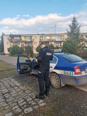 Zdjęcie przedstawia funkcjonariusza, który stoi przy radiowozie oznakowanym w pobliżu budynku wielorodzinnego. Policjant wykonuje czynności związane z telefonowaniem do osób przebywających na kwarantannie