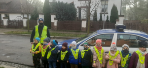 Zdjęcie grupowe, na którym widać policjanta wraz z grupą przedszkolaków ubranych w kamizelki odblaskowe. Wszyscy stoją przed przedszkolem na tle oznakowanego radiowozu.