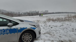 Na pierwszym planie znajduje się przód oznakowanego radiowozu. Na drugim planie dwóch umundurowanych policjantów idzie przez ośnieżone pole w poszukiwaniu za zaginionym mężczyzną.