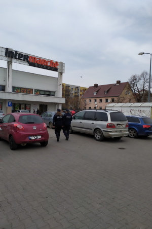 Zdjęcie przedstawia dwie policjantki, które idą przez parking pomiędzy pojazdami do marketu na kontrole