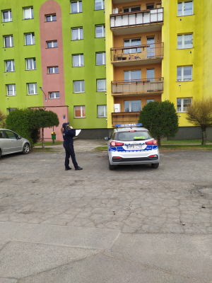 Zdjęcie przedstawia policjantkę, która stoi z telefonem pod blokiem mieszkalnym i sprawdza kwarantanny