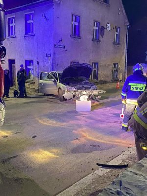 Zdjęcie przedstawia rozbity samochód osobowy marki Ford koloru srebrnego. Samochód stoi w poprzek drogi blisko budynku wielorodzinnego.
Po lewej stronie stoją ludzie przyglądający się zdarzeniu. Po prawej stronie stoi strażak z tarczą do kierowania ruchem