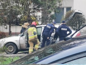 Zdjęcie przedstawia srebrny samochód osobowy Ford Mondeo, który ma otwarte drzwi kierowcy oraz bagażnik. Przy pojeździe stoi kobieta o blond włosach w czarnej kurtce, dwóch funkcjonariuszy policji oraz strażak straży pożarnej