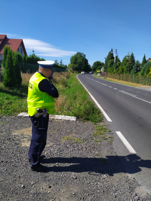 Zdjęcie przedstawia umundurowanego policjanta ruchu drogowego, który stoi przy drodze i wykonuje pomiar nadjeżdżającego z naprzeciwka pojazdu osobowego