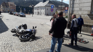 Na zdjęciu widać pięć osób, które stoją na drodze. Pomiędzy nimi leży przewrócony motocykl oraz rower- pojazdy biorące udział w kolizji. Na drugim planie widać ratusz miasta, zaparkowane pojazdy oraz miejscowe zabudowania