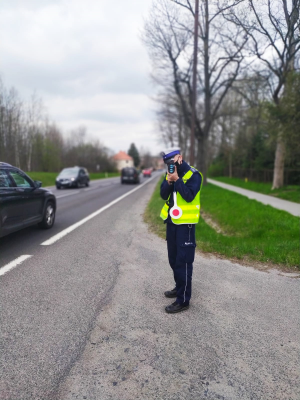 Zdjęcie przedstawia policjanta ruchu drogowego w odblaskowej kamizelce, który stoi przy drodze i wykonuje pomiar prędkości