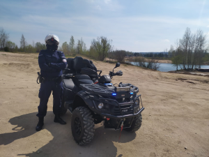 Zdjęcie przedstawia umundurowanego policjanta w kasku, który stoi przy policyjnym quadzie nieopodal zbiornika wodnego