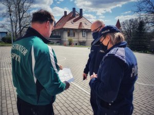 Zdjęcie przedstawia dwóch umundurowanych funkcjonariuszy policji oraz nauczyciela wfu, którzy stoją na placu przed szkołą i analizują projekt trasy rowerowej