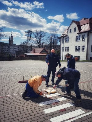 Zdjęcie przedstawia dwóch umundurowanych funkcjonariuszy policji oraz dwóch panów, którzy znajdują się na placu przed szkołą i malują przejście dla pieszych