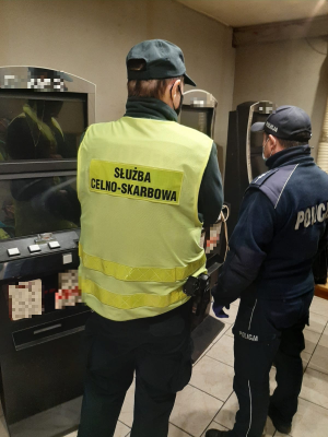 Zdjęcie przedstawia funkcjonariusza służby celno- skarbowej oraz policjanta, którzy stoją przed trzema automatami do gier hazardowych