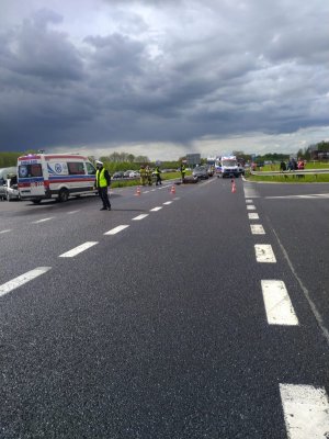 Zdjęcie przedstawia miejsce zdarzenia drogowego. Policjant ruchu drogowego kieruje ruchem, stoją dwie karetki, na asfalcie leży motocykl, bok stoi dwóch strażaków, dwóch policjantów wykonuje czynności przy motocyklu, za barierką stoi czterech  obserwujących