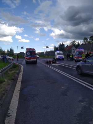 Zdjęcie przedstawia miejsce zdarzenia drogowego. Stoją trzy karetki, na asfalcie leży motocykl, obok stoją strażacy, policjanci i uczestnicy zdarzenia drogowego