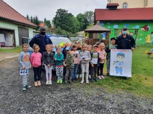 Zdjęcie przedstawia grupę przedszkolaków i dwóch policjantów którzy grupowo stoją na dworze. Jeden z policjantów trzyma plakat z wizerunkiem narysowanego policjanta