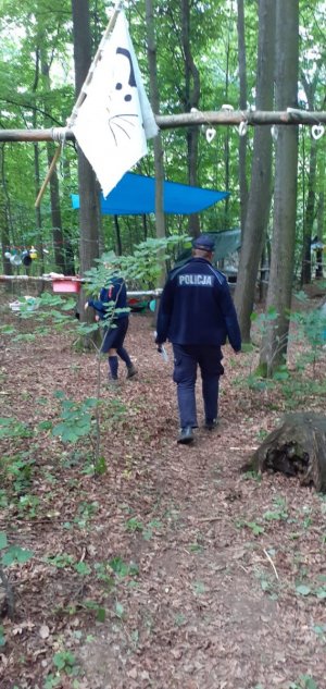 Zdjęcie przedstawia obozowisko w lesie. Na pierwszym planie białą flagę, dalej widać policjanta, który sprawdza bezpieczeństwo