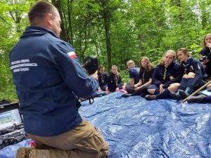 Zdjęcie przedstawia policjanta, który klęczy i w rękach trzyma kamerę noktowizyjną, którą pokazuje siedzącym naprzeciwko skautkom