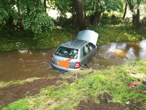 Zdjęcie przedstawia srebrną osobową skodę, która znajduje się w potoku. Pojazd ma podniesioną maskę