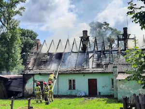 Zdjęcie przedstawia zielony dom, który ma spalony dach. Nad budynkiem unosi się dym. Strażacy wchodzą po drabinie na pozostałości dachu