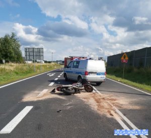 Zdjęcie przedstawia motocykl, który leży na środku drogi. Za nim stoi radiowóz oraz wóz strażacki