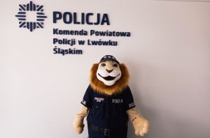 Zdjęcie przedstawia maskotkę, czyli lwa w stroju policjanta na tle napisu Policja Komenda Powiatowa Policji w Lwówku Śląskim