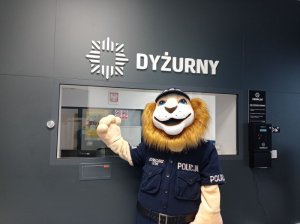 Zdjęcie przedstawia maskotkę, czyli lwa w stroju policjanta przy okienku Dyżurnego