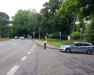 Policjant stoi przy drodze i obserwuje ruch do szkoły. Koło niego stoi radiowóz