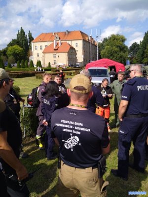 Grupa ludzi stoi na trawie podczas omówienia działań. W tle duży budynek